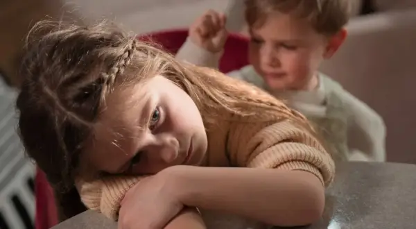 Smutna dziewczynka z warkoczem opiera głowę na rękach. Czy dziecko pamięta traumę? Za nią widać zamazanego chłopca.