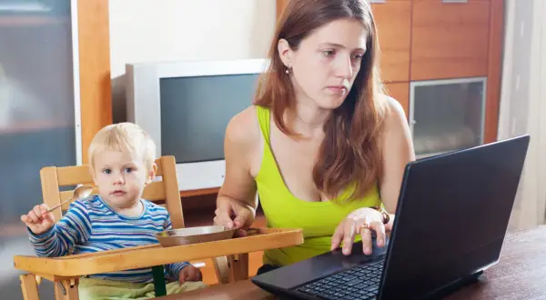Matka z dzieckiem przy stole, zastanawia się, ile trwa urlop macierzyński, pracując na laptopie.