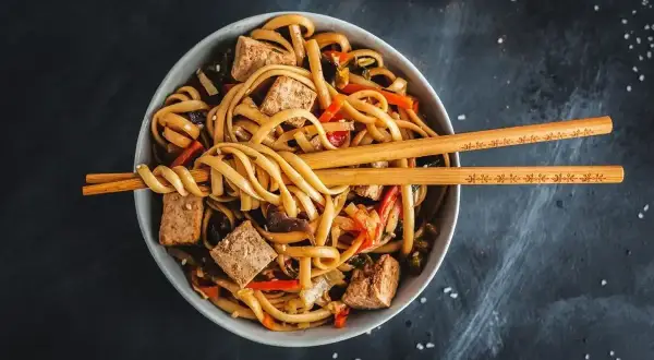 Miska z makaronem udon, tofu i warzywami, przykryta pałeczkami.