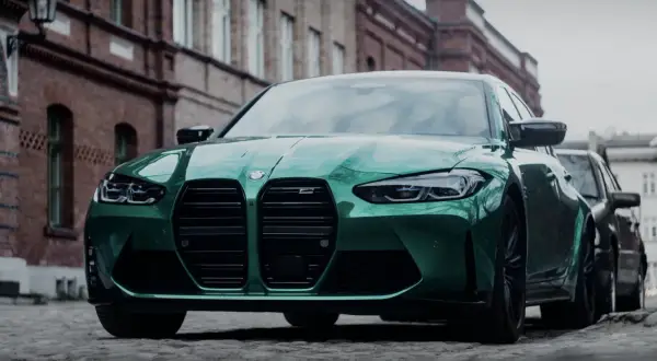 Zielony BMW M3 Competition z charakterystycznym grillem, zaparkowany na brukowanej ulicy przed ceglanymi budynkami.