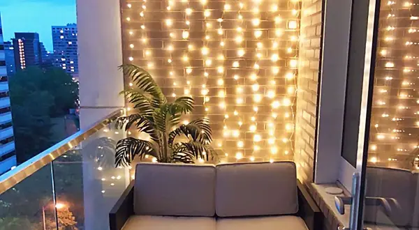 Przytulny balkon w bloku z sofą i palmą, ozdobiony ciepłym oświetleniem.