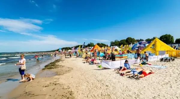 Słoneczna plaża w Ustce pełna ludzi, kolorowych namiotów i flag. Wiele atrakcji dla każdego.