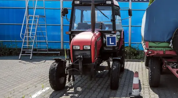 Czerwony traktor z literą "L" (kategoria t prawo jazdy) stoi obok przyczepy. W tle niebieska ściana i drabina.