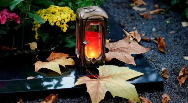 Grablicht auf einem Grabstein, umgeben von Herbstlaub und Blumen. Ein stiller Gruß zu Allerheiligen katholisch.