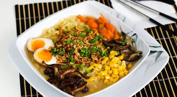 Miseczka aromatycznego tantanmen z makaronem, jajkiem na pół, kukurydzą, marchewką i grzybami shiitake, posypana szczypiorkiem.