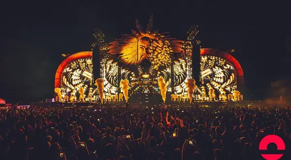 Nocny hardstyle festival z ogromną sceną i tłumem ludzi. W tle świecąca, złota głowa w koronie.