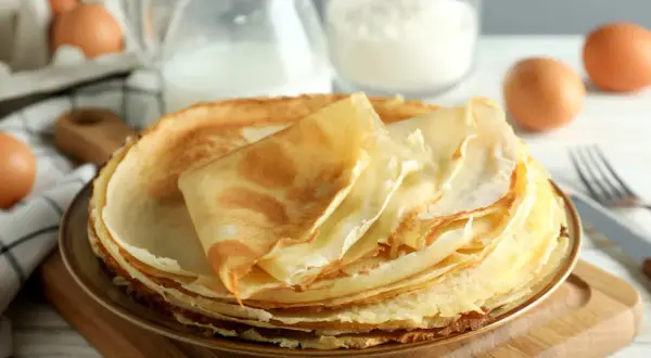 Pila di crepes dorate, pronte per essere farcite. Ingredienti per una ricetta crepes dolci perfetta: uova, latte e farina sullo sfondo.