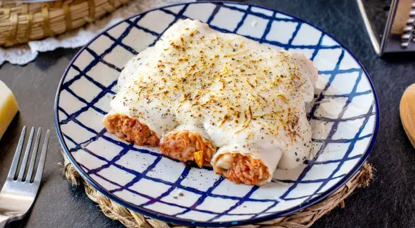 Canelones de atún y huevo gratinados, cubiertos de bechamel y queso, servidos en plato a cuadros.
