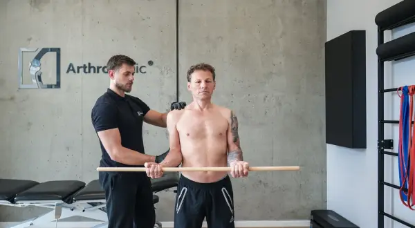 Fizjoterapeuta pomaga pacjentowi w ćwiczeniach rehabilitacja barku po operacji. Mężczyzna trzyma kij, wykonując ruchy ramionami.