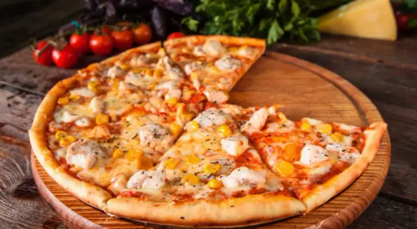 Pyszna pizza z kurczakiem i kukurydzą, podana na drewnianej desce. Obok świeże pomidorki i zioła.