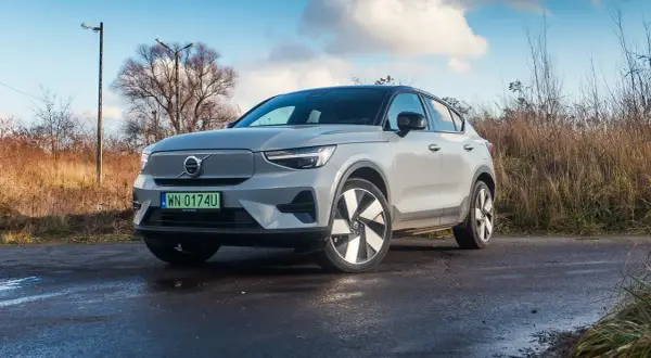 Szary Volvo C40 na mokrej drodze, otoczony suchą trawą i drzewami.