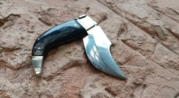 Navaja capaora de mango negro y hoja curva, lista para la acción sobre una superficie rocosa.
