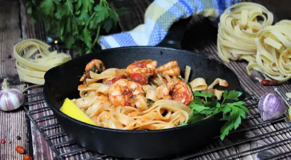 Spaghetti mit Garnelen mediterran in einer Gusseisenpfanne, garniert mit Zitrone und Petersilie. Daneben rohe Pasta, Knoblauch und Chili.