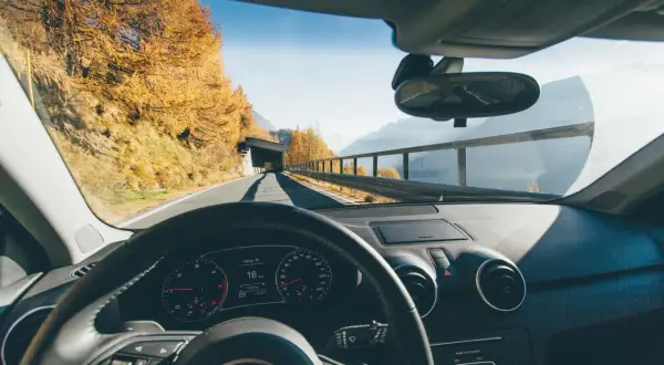 Volant d'une voiture en mouvement, tableau de bord affichant 16 km/h. Paysage d'automne en Italie, arbres dorés, montagnes et tunnel.