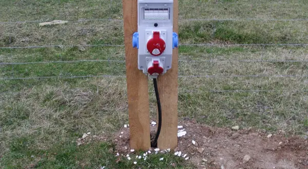 Tymczasowe gniazda elektryczne na budowie, zasilane prądem budowlanym, zamontowane na drewnianym słupku przy ogrodzeniu.