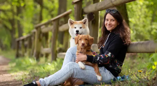 Kobieta z dwoma psami w lesie. Jeden piesek jest na jej kolanach, drugi za nim. Wskazówki, jak uspokoić sukę podczas cieczki, mogą być pomocne.