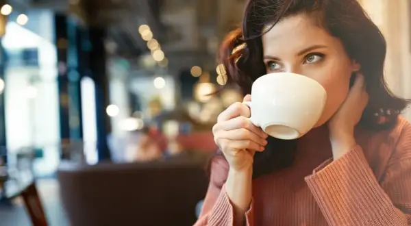 En kvinna med mörkt hår dricker kaffe ur en vit kopp. Hon har en stickad tröja och sitter på ett café. Kanske lite kaffe på tom mage för att vakna till.