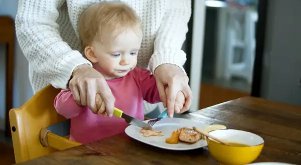 Mama pomaga dziecku kroić rybę na talerzu. To idealne pancakes dla niemowlaka, które uczy się jeść samodzielnie.