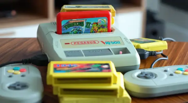 Konsola Pegasus IQ-502 z włożoną grą Teenage Mutant Ninja Turtles II i kilkoma żółtymi kartridżami oraz dwoma szarymi kontrolerami.
