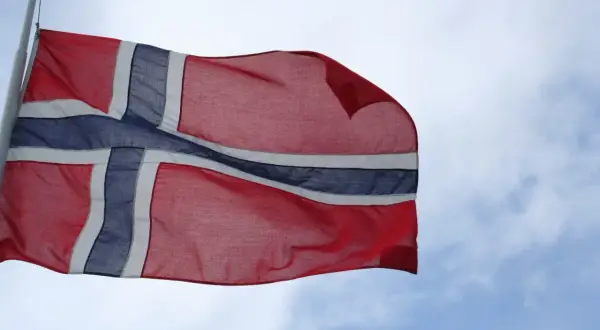 Flaga Norwegii powiewa na wietrze na tle błękitnego nieba z chmurami.