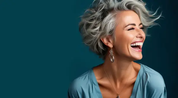 Frau mit vollem Lachen und auffälligen blau grauen Haaren, die im Wind wehen. Sie trägt einen blauen Seidenstoff und auffälligen Schmuck.