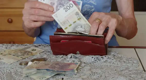 Dłonie wkładają banknot 200 zł do portfela. Na stole leżą inne banknoty i monety, co skłania do refleksji, ile wynosi średnia emerytura.