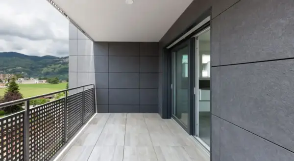 Nowoczesny balkon z jasnymi kaflemi na podłodze i szarymi płytami na ścianach. Widok na zielone wzgórza i miasto.