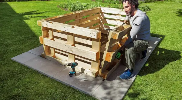 Mann baut ein schmales Hochbeet aus Paletten im Garten. Werkzeug und Schrauben liegen bereit.