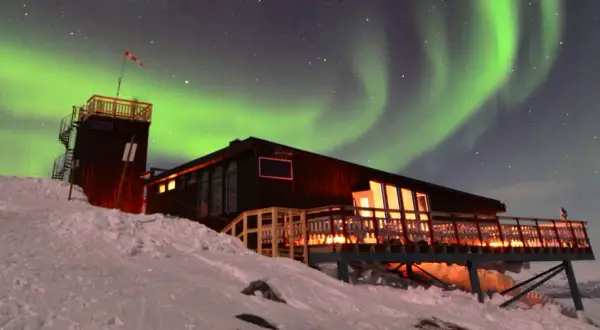 En snöig sluttning leder upp till en mysig aurora sky station. Norrskenet dansar grönt över stjärnorna.