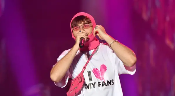 Mata raper w różowej kominiarce i okularach, z torbą na ramię i koszulką "I MY FANS", śpiewa na koncercie.