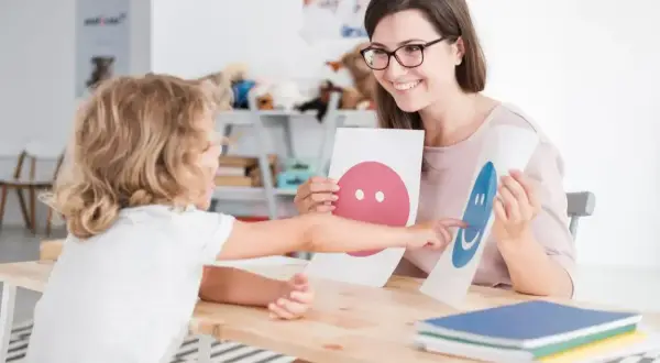Nauczycielka z uśmiechem pokazuje dziecku karty z buźkami, wspierając jego przygotowanie pedagogiczne.