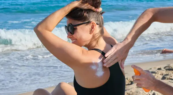 Kobieta na plaży smaruje plecy kremem z filtrem. Zastanawiasz się, ile trzeba byc na słońcu, żeby się opalić? Pamiętaj o ochronie!