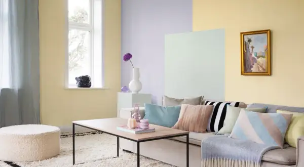 Ett vardagsrum med pastell inredning. Ljusgula, ljuslila och ljusgröna väggar skapar en mjuk atmosfär. En beigefärgad soffa är prydd med kuddar i olika pastellfärger och mönster.