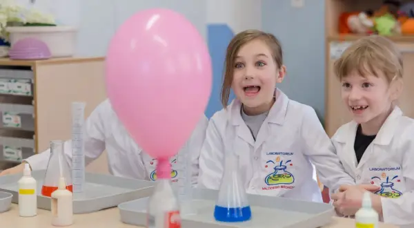 Dzieci w fartuchach laboratoryjnych z ekscytacją obserwują różowy balon podczas eksperymentów chemicznych dla dzieci.