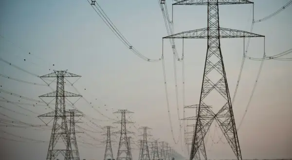 Linia wysokiego napięcia z wieżami energetycznymi, przypominającymi gigantyczne kogeneratory, ciągnie się przez pustynny krajobraz.
