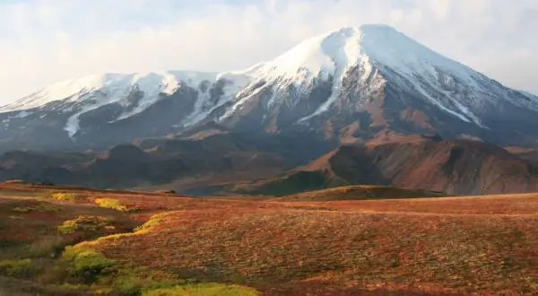 Majestätischer schneebedeckter Vulkan in einer weiten russischen Landschaft mit herbstlich gefärbten Hügeln im Vordergrund.