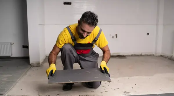 Obrero con guantes amarillos prepara un azulejo gris para pegar con Sikaflex en una obra.