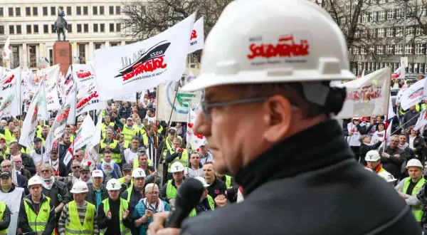 Mężczyzna w białym kasku z logo "Solidarność" przemawia do tłumu ludzi z flagami. To demonstracja związków zawodowych.