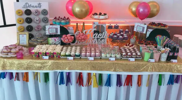 Słodki stół z donutami, babeczkami i deserami w słoiczkach, jak na wesele lata 80. Złoty obrus i kolorowe balony dodają blasku.