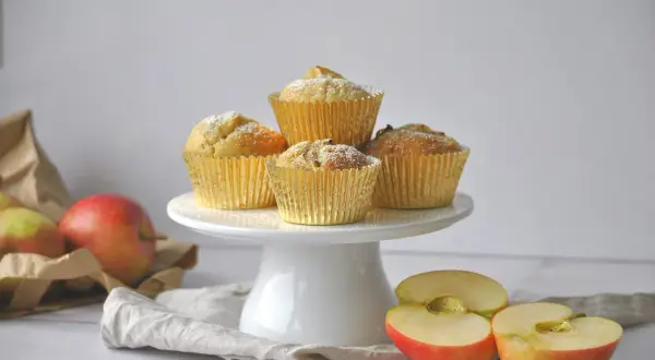 Goldene Muffins mit Puderzucker, daneben Äpfel und eine Tüte. Ein leckeres apfelmuffins rezept zum Nachbacken.