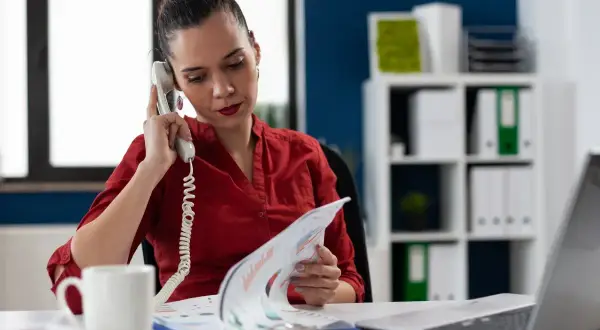 Kobieta w czerwonej bluzce prowadzi rozmowę telefoniczną, przeglądając dokumenty. Może to być element jej pracy związanej z twardą windykacją.