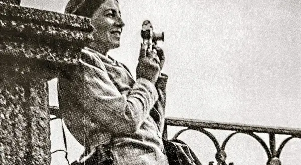 Zofia Chomętowska, z aparatem w dłoni, patrzy w dal. W tle balustrada i jasne niebo.