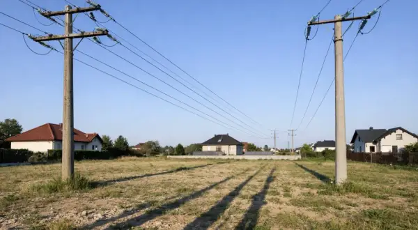 Likwidacja napowietrznej linii energetycznej. Dwa betonowe słupy z przewodami na tle domów i błękitnego nieba.
