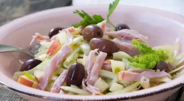 Frischer Fenchel Salat mit Apfelstücken, Schinkenstreifen und dunklen Oliven in einer rosa Schüssel.
