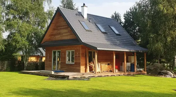 Drewniany dom kanadyjski z werandą, otoczony zielenią i drzewami. Na dachu widoczne okna dachowe i komin.
