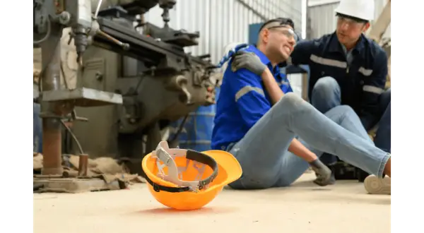 Robotnik po wypadku, kolega pomaga. Pomarańczowy kask leży obok. Sprawa dotyczy zadośćuczynienia a odszkodowania.