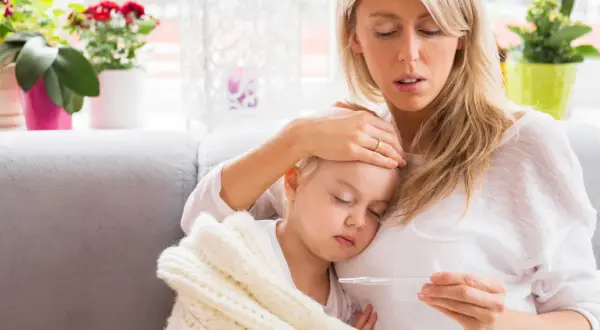 Zmartwiona mama sprawdza temperaturę chorej córki, która śpi otulona kocykiem. Myśli o podaniu Fervex dla dzieci.