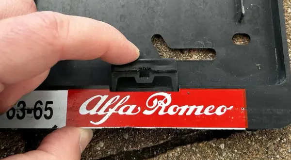 Palec naciska na zatrzask, pokazując, jak zdjąć tablice rejestracyjne z ramki Alfa Romeo.