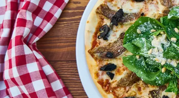 Pizza z rukolą i grzybami na białym talerzu, obok czerwono-biała kraciasta serwetka.
