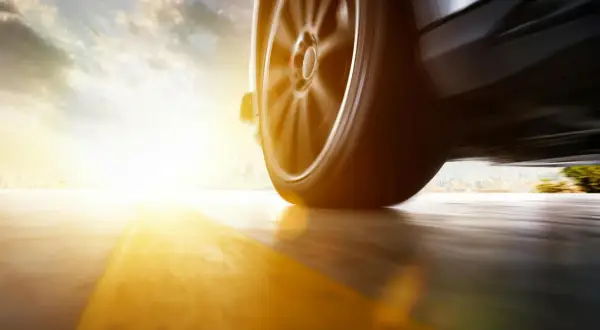 Roues d'une voiture en mouvement, le soleil rasant inonde la scène d'une lumière dorée, floutant le paysage urbain au loin.